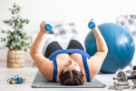 Overweight lady doing sports exercise in living room. He lies on the mat and lifts dumbbells.の写真素材