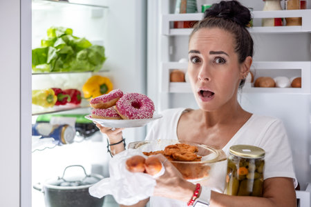 On Friday evening, a very hungry woman in pajamas chooses snacks, steaks, donuts and a can of pickles from the refrigerator.の写真素材