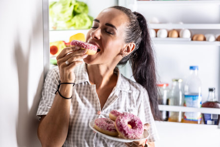 Hungry brunette in pajamas enjoys sweet donuts late at night by the open refrigerator.の写真素材