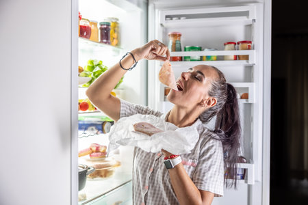 Very hungry woman in pajamas enjoying ham at night by the fridge.の写真素材