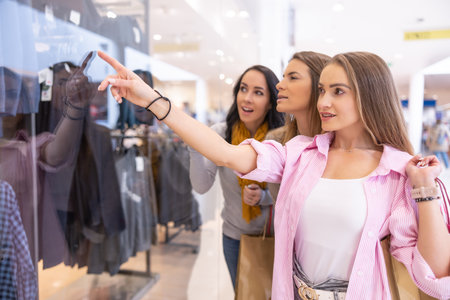 Three cheerful female friends are looking at clothes behind the glass of a store in a supermarket.の写真素材