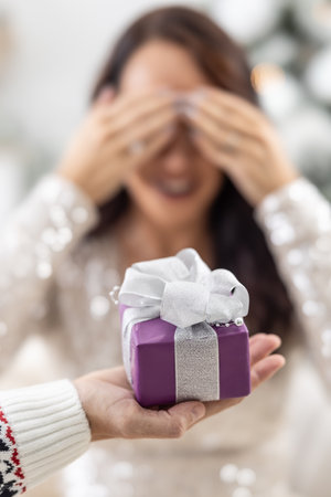 Man's hand is holding purple Xmas present with a ribbon in front of a woman closing her eyes with hands.の写真素材