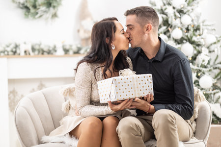 Woman and man kiss in front of the Christmas tree holding a Xmas present together.の写真素材