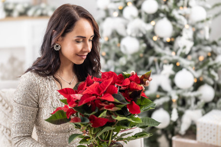 Woman smiles and smells a poinsette in a flower pot next to a Christmas tree.の写真素材