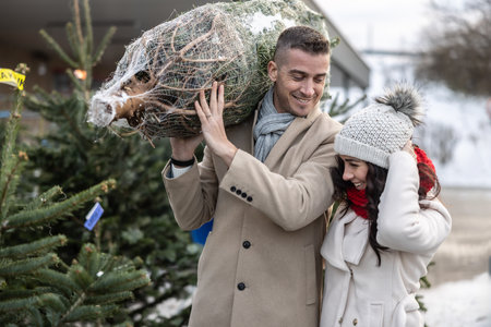 A young man carrying a wrapped Christmas tree bought at the market together with his wife. The happy couple bought a Christmas tree.の写真素材