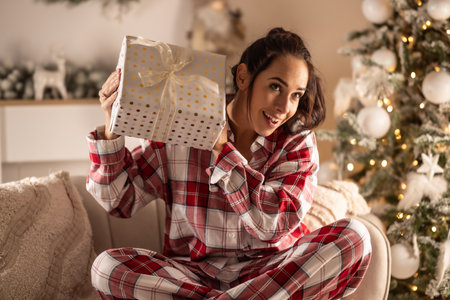 Curious cheerful woman in pajamas shakes a Christmas present and listens to what it could be, sitting next to a Christmas tree.の写真素材