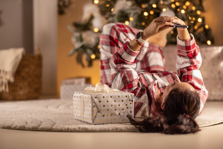 Cheerful woman in pajamas lying on carpet in living room with gift next to Christmas tree doing video chat with friends.の写真素材