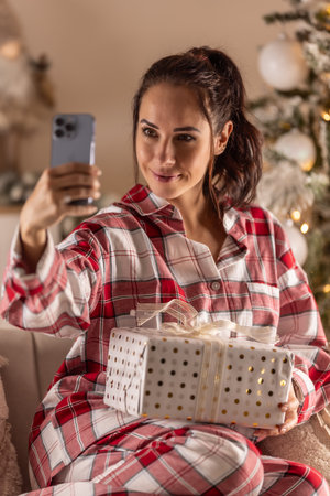 Cheerful woman in pajamas sitting on sofa with gift next to Christmas tree taking selfie.の写真素材