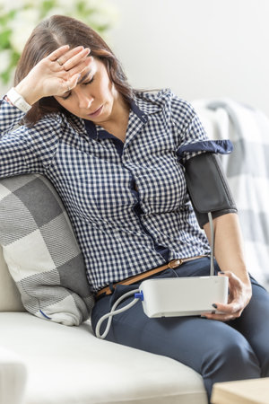 A young woman has a problem with dizziness, sits on a sofa and measures her blood pressure with a digital measuring device.の写真素材