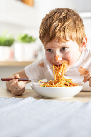 A cute little boy is eating spaghetti bolognese for lunch in the kitchen at home and is covered in ketchup.の写真素材