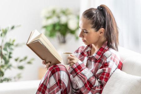 Beautiful brunette in pajamas is reading a book sitting on the sofa in the living room.の写真素材