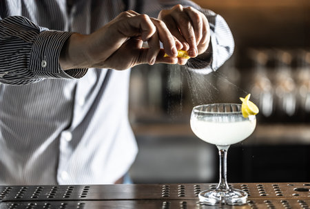 A bartender squeezes a lemon peel and squirts the juice onto a French 75 cocktail drink on the bar counter.の写真素材