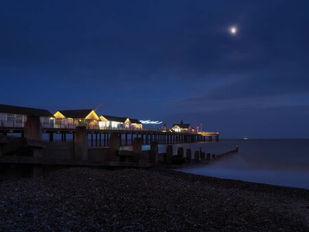 Southwold Pier with buildings and lights lit up at night and waves flowing onto a pebble beach next to a wooden groyne under a bright moon with a ship on the horizon, Suffolk, UKの写真素材