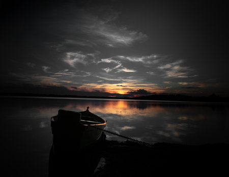 A peaceful lake at sunset with a silhouetted boat anchored on the shore, reflecting golden hues in the calm watersの写真素材