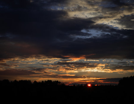 A striking sunset with deep, textured clouds casting dramatic shadows across the sky. The golden light breaking through the darkness creates a powerful contrast between light and shadow.の写真素材