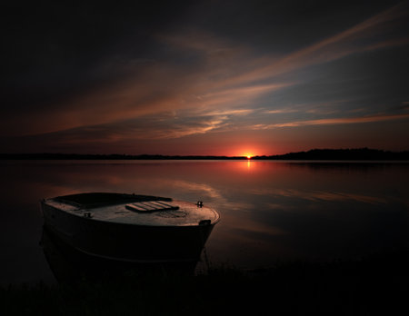 A serene yet moody sunset casts a warm glow over a calm lake, reflecting the rich hues of the fading light. A solitary boat rests near the shore, adding depth and emotion to the tranquil landscape.の写真素材