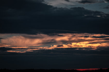A moody sunset sky with layers of dark clouds and a golden glow breaking through. The contrast between shadow and light creates a powerful, cinematic atmosphereの写真素材