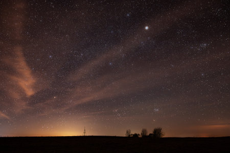 A wide-angle capture of a dazzling star-filled night sky above a silhouetted landscape and triangulation tower in Lithuania. Visible constellations and warm light pollution.の写真素材