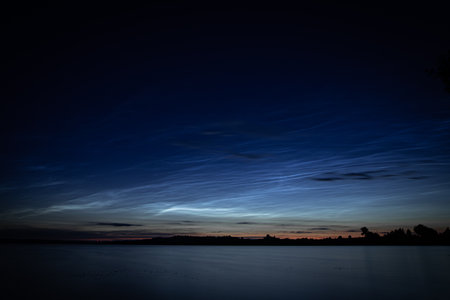 Delicate noctilucent clouds stretch across a twilight sky above a tranquil summer lake. Their shimmering bluish hues create an ethereal contrast against the fading glow of sunsetの写真素材