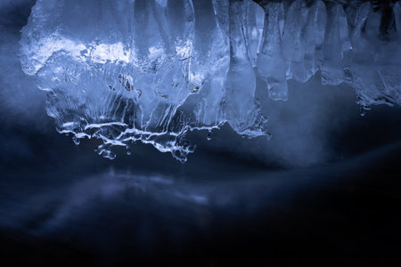Sculptural ice formations hang above a stream of dark, moving water, illuminated with cool blue tones. The organic shapes and reflective surfaces create an abstractの写真素材