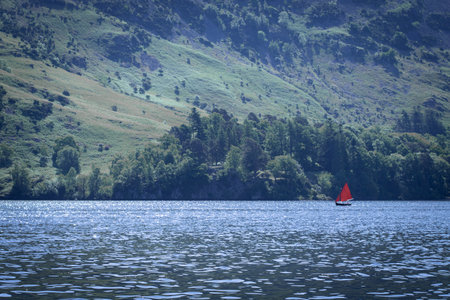 A vibrant red-sailed boat glides across a shimmering deep-blue lake, set against lush, sunlit mountain slopes. The striking contrast of the sail and water evokes calm poetry.の写真素材