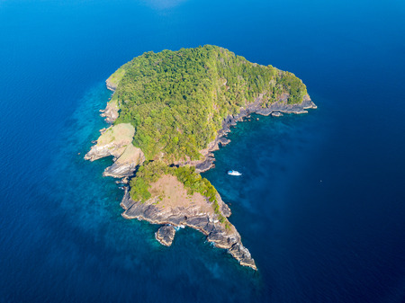 A single boat next to a remote, uninhabited tropical island covered in green jungle and surrounded by tropical coral reefの写真素材