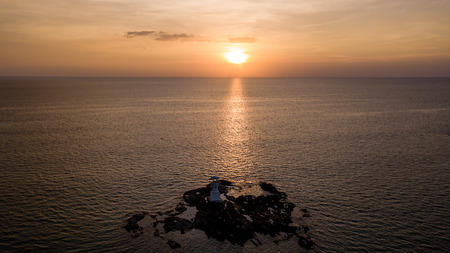 Drone view of a beautiful tropical sunset beind a small, white lighthouse located on rocks next to...
