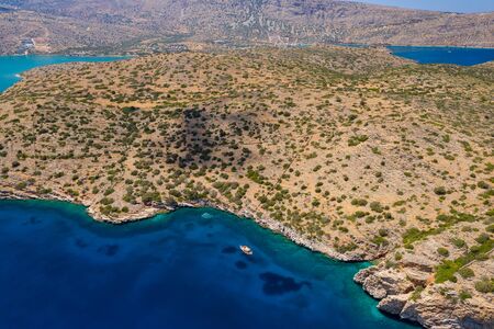 Aerial drone view of boats in clear, blue water next to a dry, yellow summer coastline (Crete,...