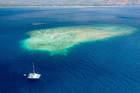 Aerial view of a sailboat next to a large, offshore tropical coral reef