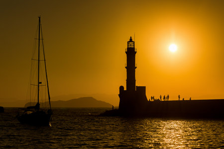 Silhouette of a lighthouse and small yacht during a beautiful golden sunset on a Greek island in summerの写真素材