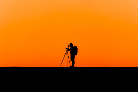 Silhouette of a lone photographer with tripod taking an image during a beautiful orange sunsetの写真素材