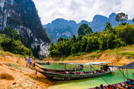 KHAO SOK, THAILAND - APRIL 02 2024:- Wooden longtail boats on the banks of a jungle lake surrounded by lush rainforest and towering limestone cliffsのeditorial素材