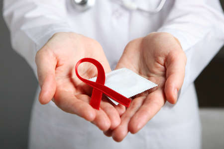 Red ribbon symbolizing AIDS and condoms in the hands of a doctor in a white coatの写真素材