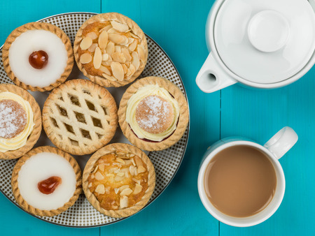 Plate of Assorted Individual Cakes or Tarts With a Pot of Tea Against a Blue Backgroundの写真素材