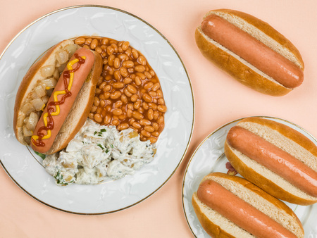 Hot Dog in a Bun with Baked Beans and Potato Salad Against a Pale Orange Backgroundの写真素材