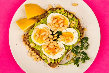 Sliced Boiled Egg on Crushed Avocado With Pine Nuts And Rye Bread Open Faced Sandwich Against A Pink...