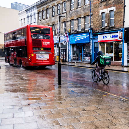 London UK, March 26 2021, Deliveroo Home Dleivery Cyclist Following A Red Double Decker Public...