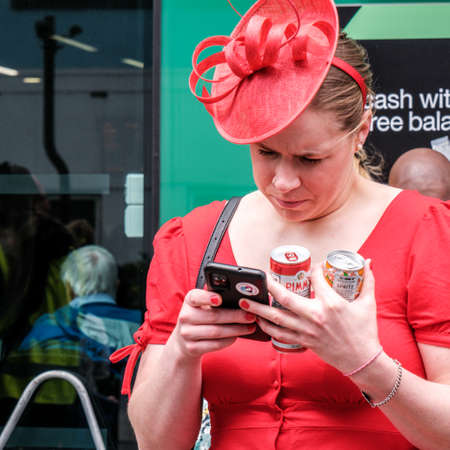 Epsom Surrey, London UK, June 04 2022, One Woman Wearing Red Dress And Fascinator Using Mobile Phone And Holding Cans Of Pimms Alcoholic Drinkのeditorial素材
