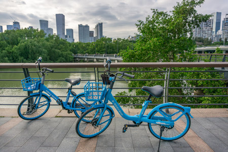 Shenzhen, China - November 12, 2025: Two blue shared public bicycles parked on pedestrian bridge waterfront with river, green trees and modern city skyscrapers skyline in background on cloudy day.のeditorial素材