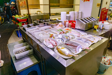 Shenzhen, China - November 12, 2025: Street restaurant display counter with variety of fresh fish and seafood wrapped in plastic on plates including yellow croaker and squid ready for cooking.のeditorial素材