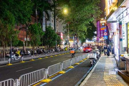 Shenzhen, China - November 12, 2025: Busy city street at night with densely parked mopeds, moving cars, pedestrians and food delivery riders under bright neon signs and streetlights.のeditorial素材