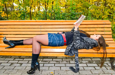 young brunette woman in miniskirt reading book on bench in autumnal parkの写真素材