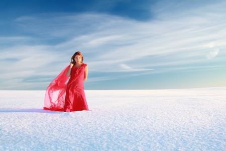 Young woman in red dress with translucent shawl in the snow desert under blue skyの写真素材