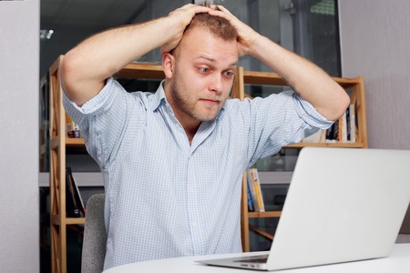 young businessman at his desk looking fearfully  looking at a laptop in his bright office