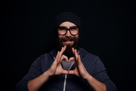 Handsome male big beard in glasses and hat, studio shot on black background, man shows heart fingersの写真素材