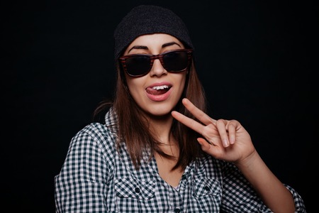 Hipster teenage girl with beanie hat and plaid shirt posing making confident facial expression, smiling, showing two fingers against the black wall background. Modern young people.の写真素材