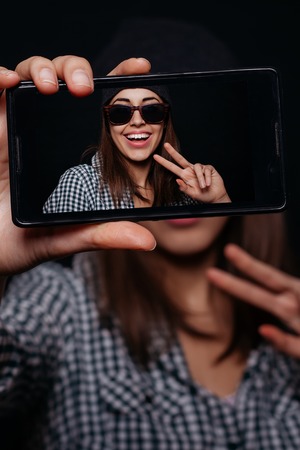 Hipster cool girl taking picture on smartphone self-portrait, screen view, snapshot studio on a black backgroundの写真素材
