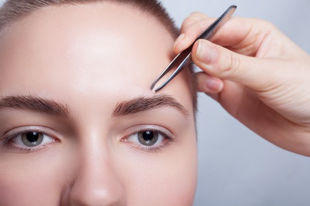 Young woman with short hair plucking eyebrows with tweezers close up, studio shot. on a light background. beauty shot .Closeup part of face, woman plucking eyebrows depilating with tweezers.の写真素材