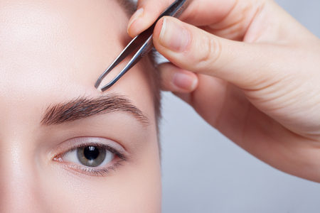 Young woman with short hair plucking eyebrows with tweezers close up, studio shot. on a light background. beauty shot .Closeup part of face, woman plucking eyebrows depilating with tweezers.の写真素材