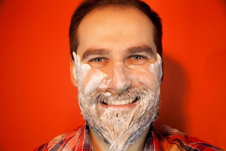 Handsome man with shaving foam on his face and razor, smiling, happy on a red background, color photoの写真素材